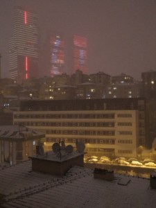 Nocturnal scene of city buildings under snow, loomed over by towers displaying the crescent and star of the Turkish flag in colored lights