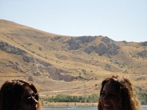 Two women talking on shipboard; behind them, brown hills, with green trees near the shore