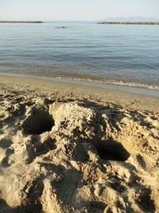 Two holes in the sand of a beach; calm sea beyond (one person in it), breakwaters, and blue sky