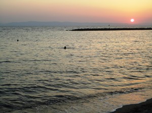 Mostly a calm sea, with heads of two swimmers; behind, a strip of pink sky with setting sun
