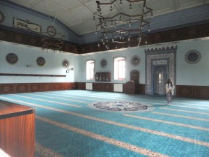 The ceiling of white panels is low for the space. The carpet on the floor is turquose with lighter strips, which are parallel to the far wall, in which are two windows and the mihrab