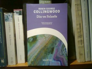 Book showing its front cover, reading “Robin George / Collingwood / Din ve Felsefe / İngilizce aslından çeviren: / Fulya Kılınçarslan / akademim,” resting against the spines of other books on a shelf
