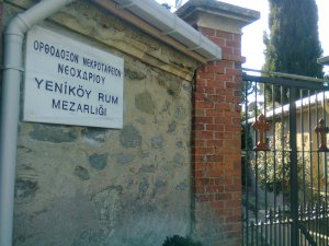 Stone wall next to iron gate with two Byzantine cross-crosslets. Sign on wall is engraved with words in Greek and Turkish: “ΟΡΘΟΔΟΞΟΝ ΝΕΚΡΟΤΑΦΕΙΟΝ / ΝΕΟΧΩΡΙΟΥ / YENİKÖY RUM / MEZARLIĞI”