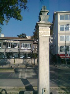 Square stone column with faucet at the bottom and Ottoman writing in Arabic script at the top, below the capital; road, buildings, and trees behind