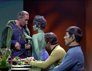 Thumbs in his belt, Garth looks down at Marta, who returns the look, her hand on his chest; seated, forearms on table, Kirk looks on, while Spock, arms crossed, looks into the distance