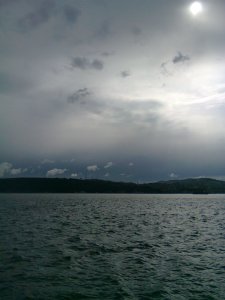 Sun through clouds of many shades; below a strip of dark hills, the sea reflects the light of the sky