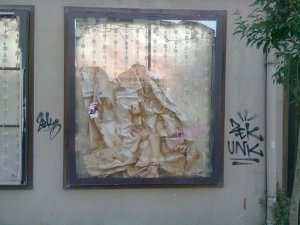 Crumpled paper on shiny ground behind glass within a dark frame on an exterior wall; graffiti tags on either side; right side of similar frame on the left, small tree on the right