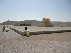 All of yellowish stone, a box with roughly half-cylindrical roof sits on a stepped pyramid. The whole thing is on a large raised square, covered with gravel, as are the surroundings. A few tourists read the plaques just off the square. At the near corner is a spotlight and a trashcan. In the distance, trees, and behind them, mountains, dotted with a few trees