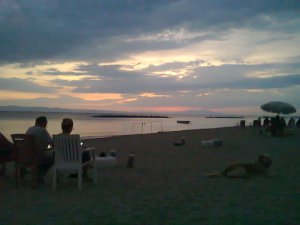Above, a sky partly filled with dark clouds, between which are patches of white and pink from a sun recently set behind distant mountains; below, separated by a triangle of sea, is a beach where, sitting on chairs facing west, two figures, male and female, lower their heads to contemplate glowing screens