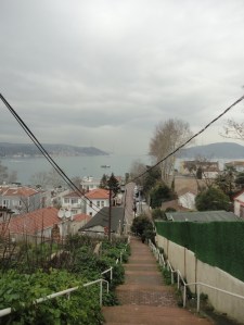 The railings beside a long descending stairway, and two cables above, converge on a village by water on a cloudy day