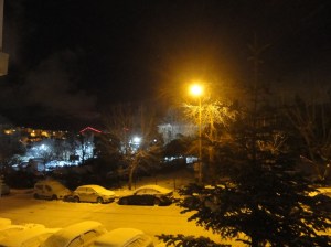 At night, a streetlight illuminates a street lined with cars, all covered with snow. In the distance, the red cable