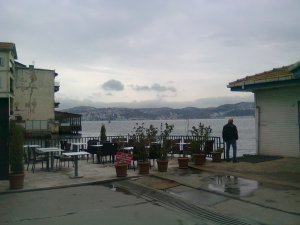 Cafe tables with sea beyond (and beyond that, land). Houses standing right on the sea are on the left; on the right, a man next to a small building looks out to sea