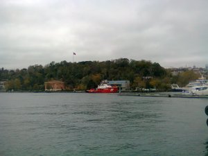 Between water and cloudy sky, trees on a hillside, with two boxy buildings at the base, and in front of them, a ship with red hull; more ships docked at the right