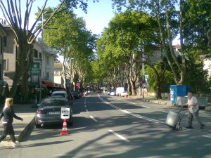 Trees in leaf line a broad street into the distance. In the foreground, a women on the left sets out to cross; on the right; a man with a barrel is halfway across in the other direction