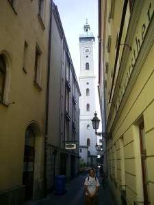 A figure poses in the narrow alleyway between two buildings; beyond, a thin square tower