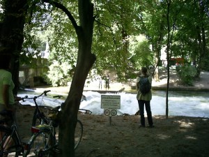 Separate figures, one with bicycle, stand beneath trees watching surfers near a sign saying, “Surfen und Baden verboten”