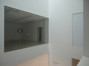 Two blank walls meet. Much of the left wall is a plate of glass. The room seen through the glass is empty, but for two or three small chairs and a video monitor in the corner showing the face of a man with a mustache. A passage to another room is partially covered by a curtain