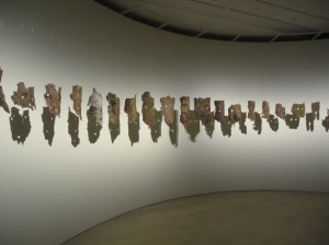 Against a concave white wall, a line of rough masks, two eye-holes each, made of tree bark