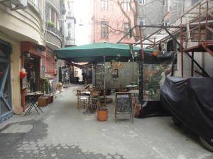 Tables and chairs beneath a big umbrella between buildings. There is no passage for cars. A chalkboard reads, “MAWAA // coffee // food”