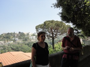 Photo: Two figures in shadow; behind them, a roof, an umbrella pine, and a hill of trees and houses