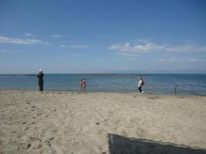 Seen from a beach, two fully clothed woman stand against the sea, holding up mobiles, one facing the sea obliquely, the other walking along the shore; in the sea, a woman and man stand together, in bikini and trunks respectively. Beyond them all, two breakwaters in line, parallel to the shore, made of rubble
