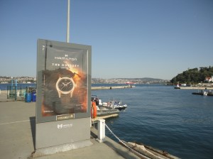 Against the backdrop of a harbor, a poster advertises an Odyssey-themed wristwatch